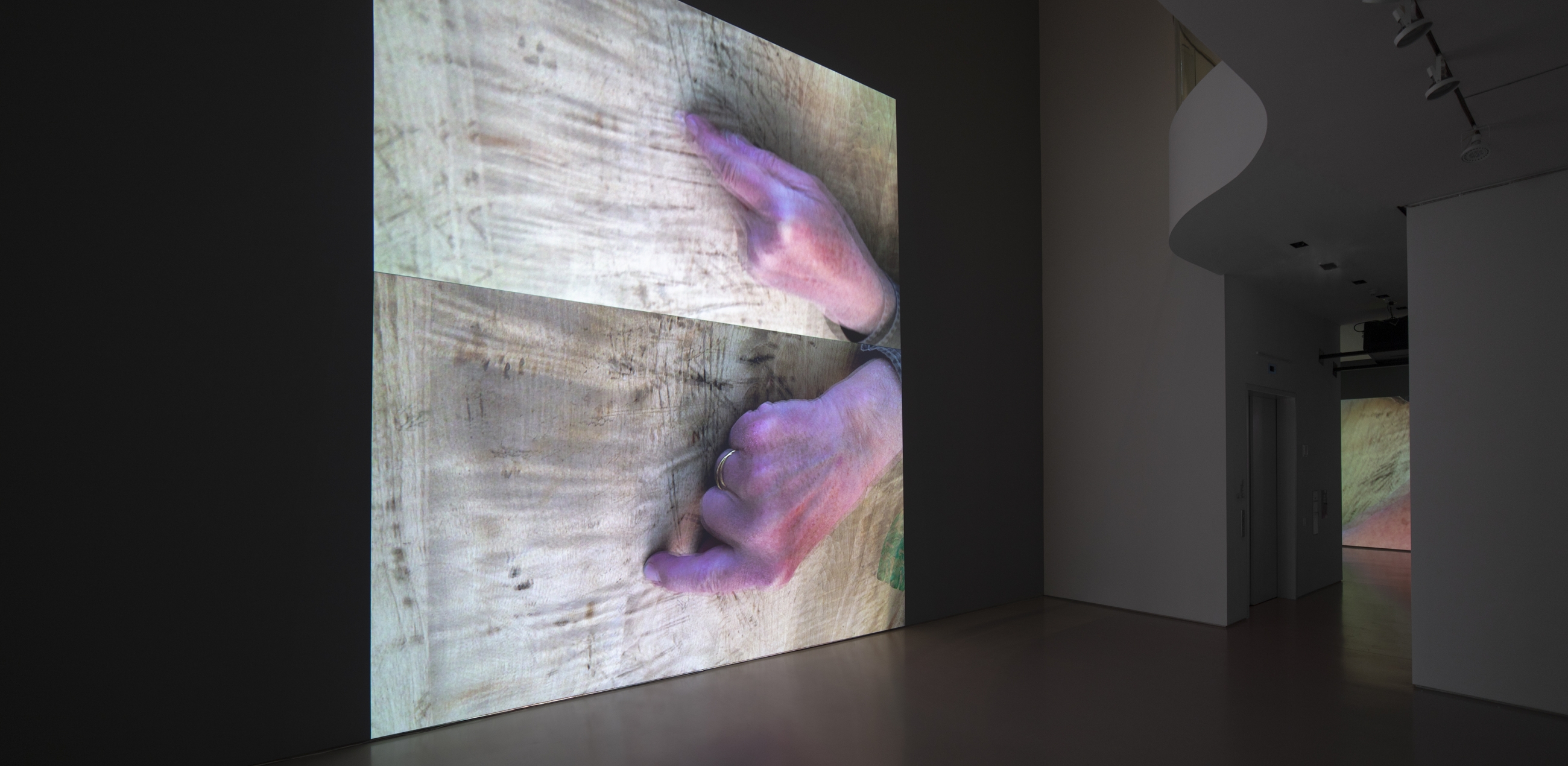 installation view of two video projections on gallery wall depicting the artist's hands on a wooden table