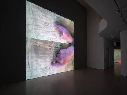 installation view of two video projections showing large hands over a wooden table