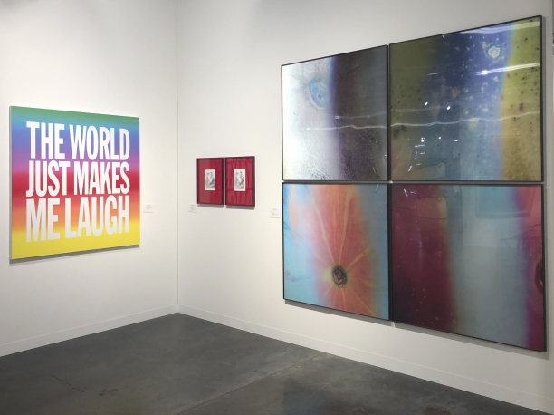 Three artworks hang in a gallery. A rainbow painting by John Giorno reads "THE WORLD JUST MAKES ME LAUGH". There are holographic, colorful abstract photographs by Jitish Kallat and a red diptych by Andrew Sendor.