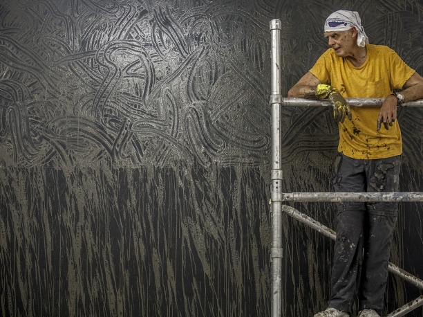 Richard Long standing on scaffolding in front of a mud artwork on the wall