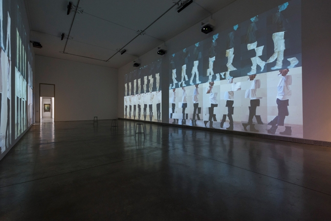 installation view of a darkened gallery with large-scale projections of two rows of a male figure stacked and segmented