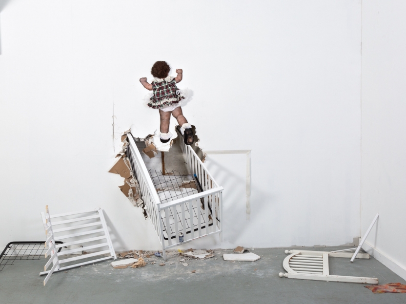 sculpture of a toddler in a frilly dress and her crib bursting through a white wall with broken crib pieces on the floor
