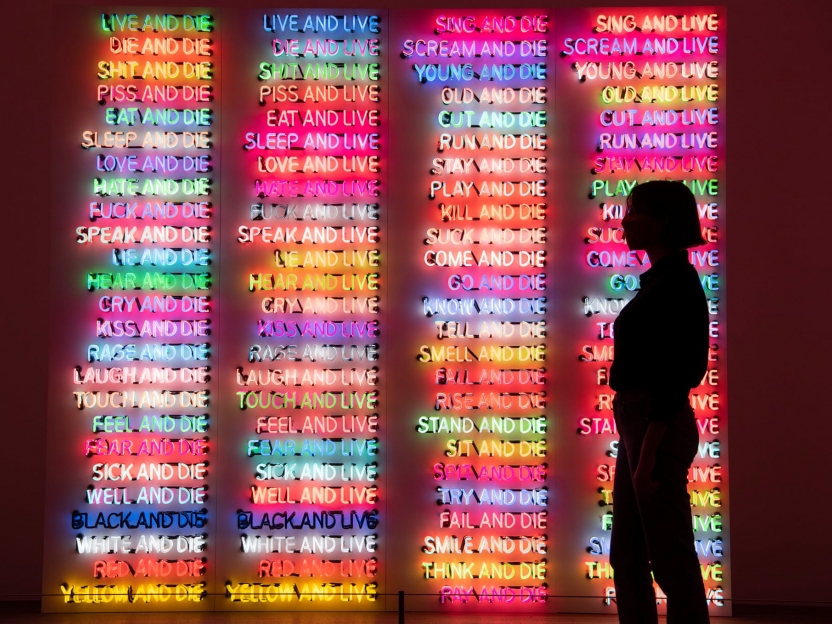 a figure in silhouette stands in front of a large-scale artwork comprised of short phrases in glowing neon