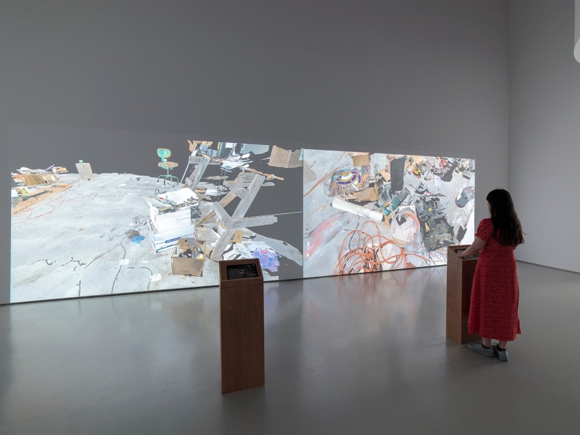 a woman stands at one of two pedestals in a gallery in front of two large-scale projections showing an artist's messy studio