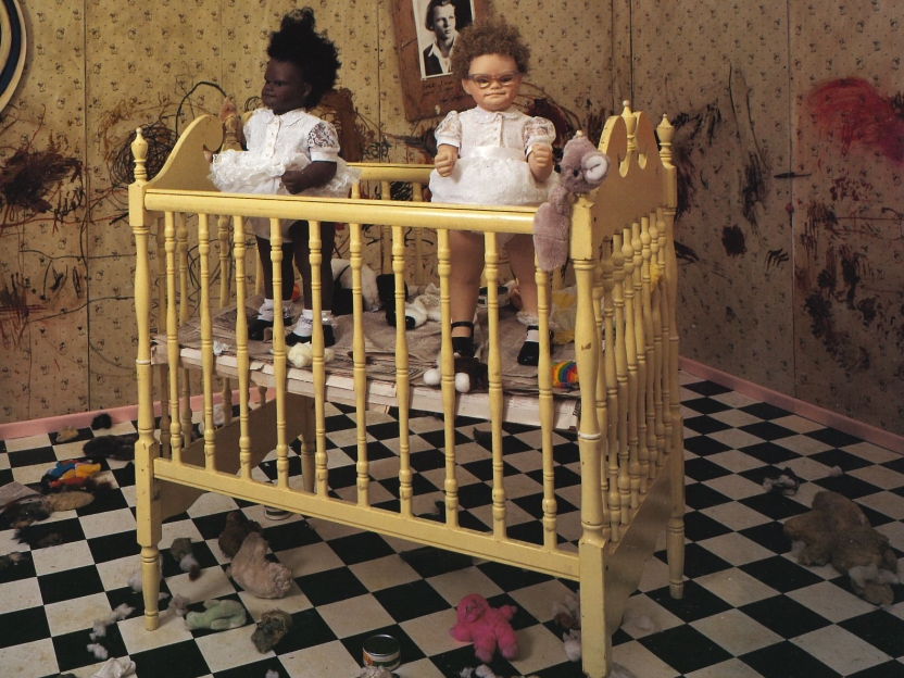 sculptures of two dolls in frilly dresses stand in a crib in a room strewn with toys