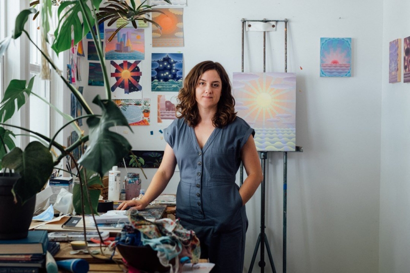 artist Amy Lincoln stands in her studio with an easel and artwork on the wall behind her